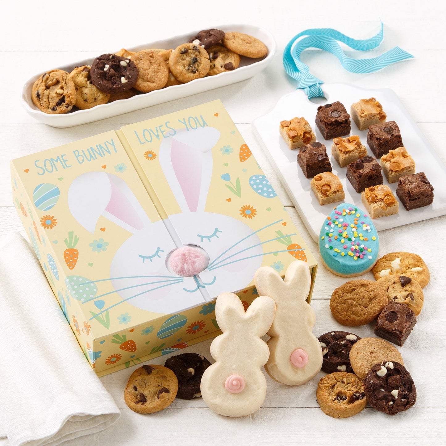 A bunny gift box surrounded by an assortment of nibblers, brownie bites, a frosted egg cookie and frosted bunny cookies